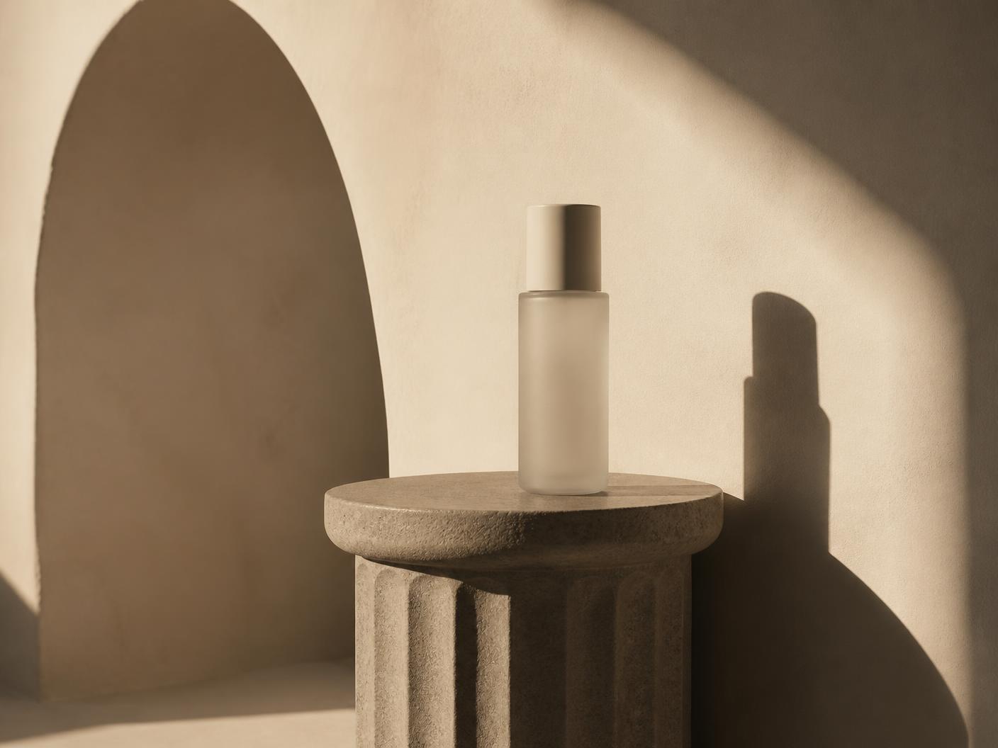 Warm ivory plaster wall with a soft arch shadow and a fluted stone pedestal holding an unbranded skincare bottle